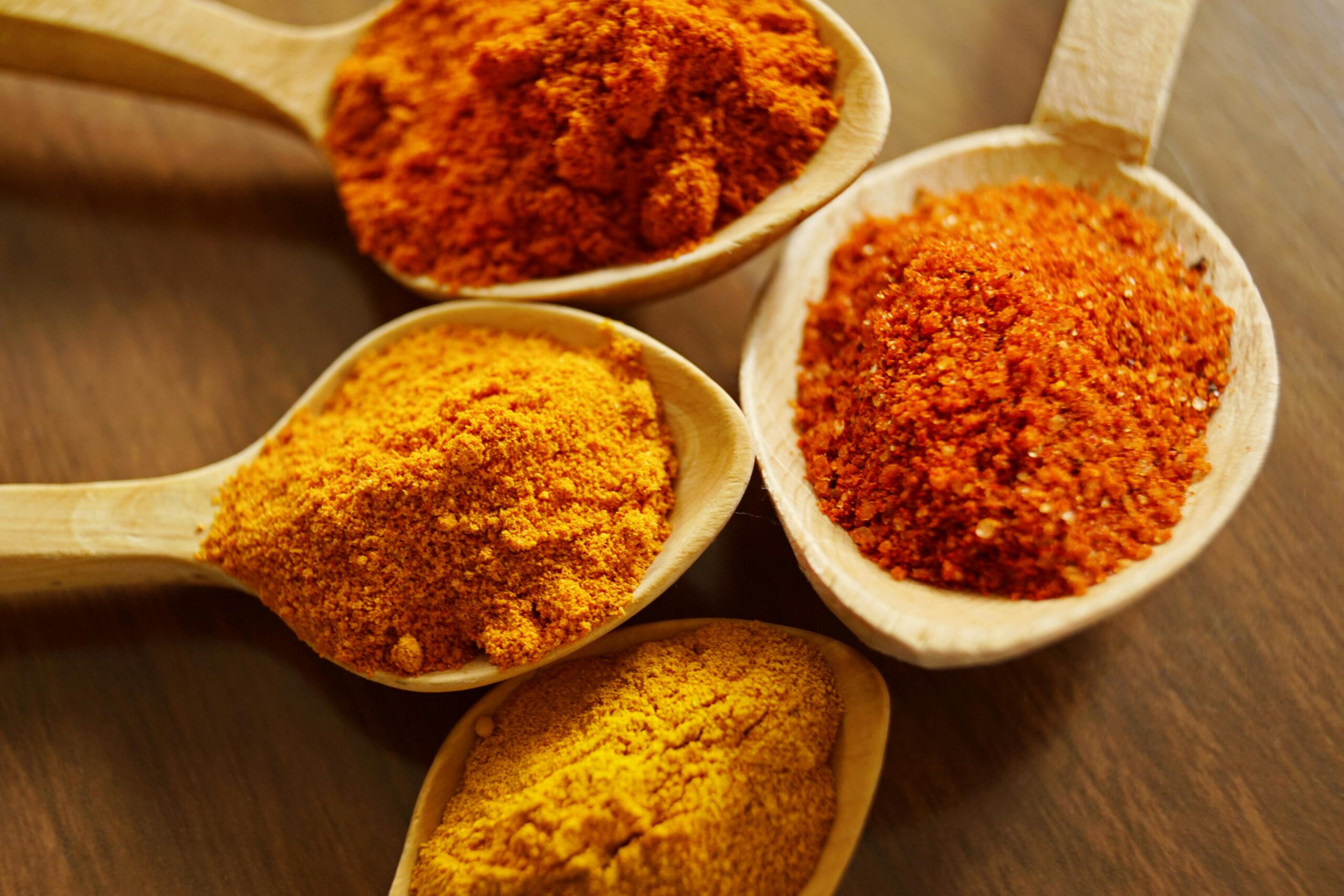 A close-up image of four spoons with different spices on a wooden surface