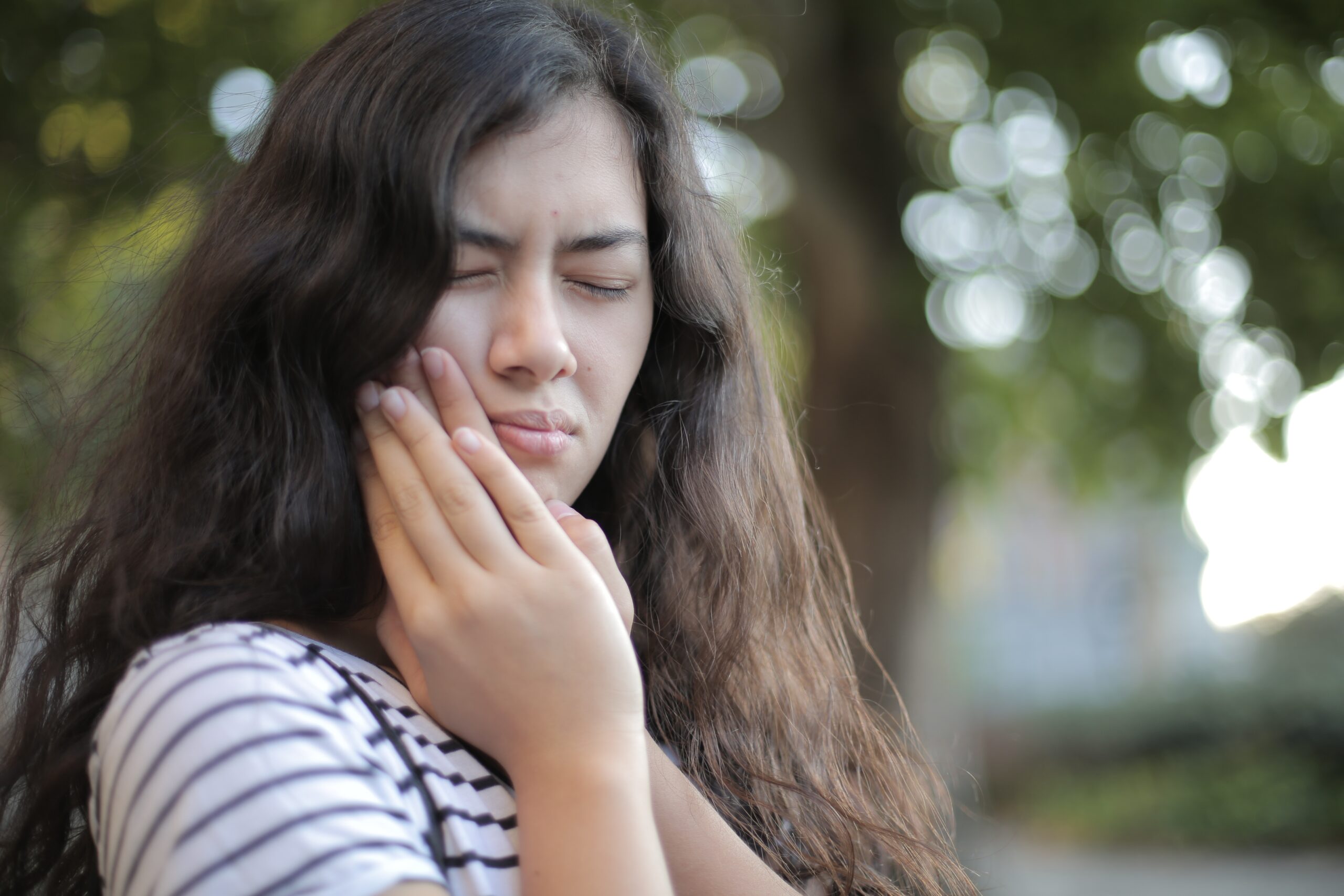 Woman holding her jaw in pain, indicative of mouth discomfort, with a blurred outdoor background
