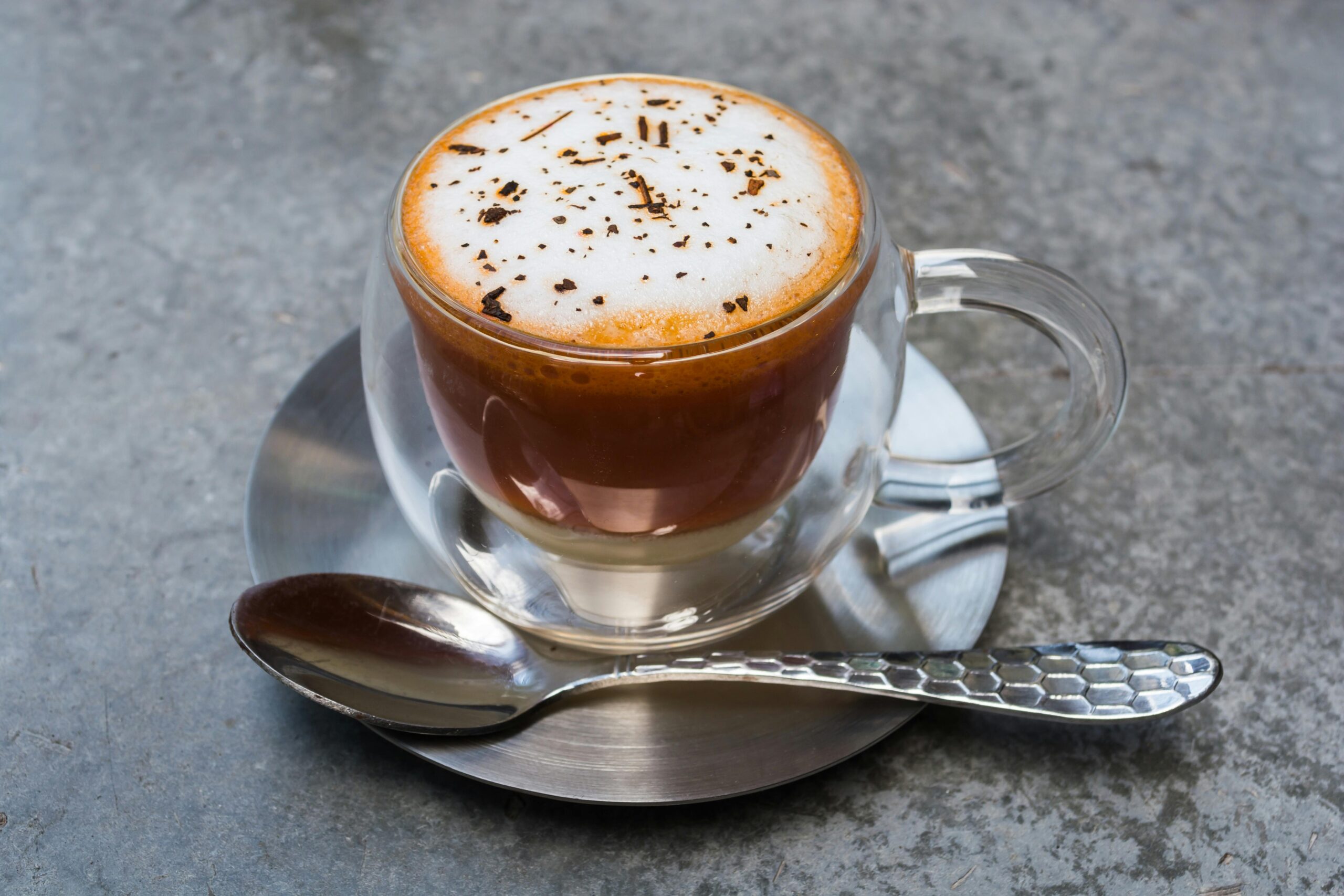 A clear glass cup of coffee resting on a saucer with a metal spoon, ready to be enjoyed.