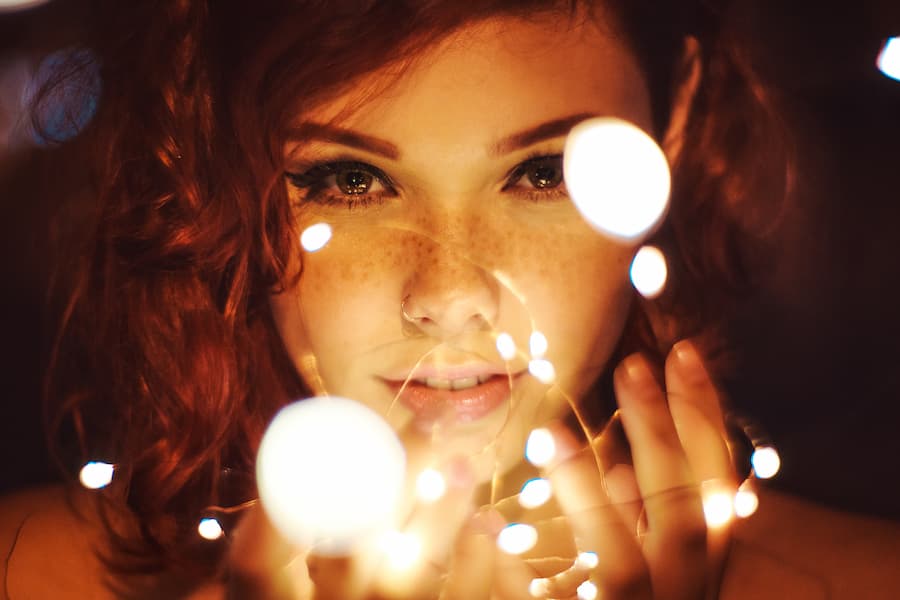 A young woman with freckles looking directly into the camera with a soulful expression, holding glowing fairy lights that illuminate her face in a warm, bokeh light effect.