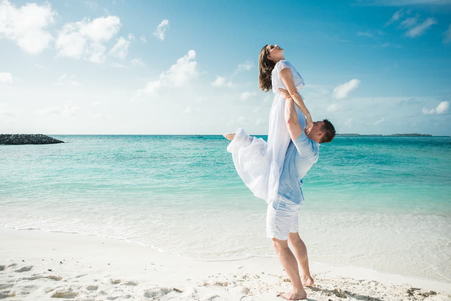 A man lifting a woman in a white dress on a beautiful sunny beach with turquoise water, capturing a moment of pure joy and freedom.