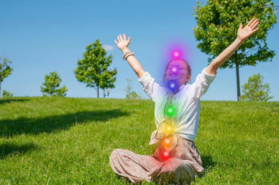 A happy woman sitting in a lotus position on green grass, arms raised in joy, with colorful chakra symbols glowing on her body to represent spiritual alignment and finding one's purpose.